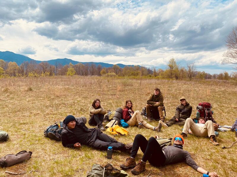 A group of people are resting in a field. Some are lying down, while others are sitting. They appear to be taking a break from hiking, as they have backpacks and are surrounded by nature. The sky is cloudy, and there are mountains in the distance. The overall scene suggests a peaceful and relaxing moment during an outdoor adventure.